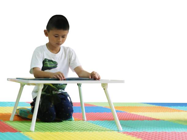 istock A young Asian boy is sitting on a colorful floor, learning / studying / reading a book on a table.  Selective focus at the boy, isolated on white background and blurred logos. 946592844