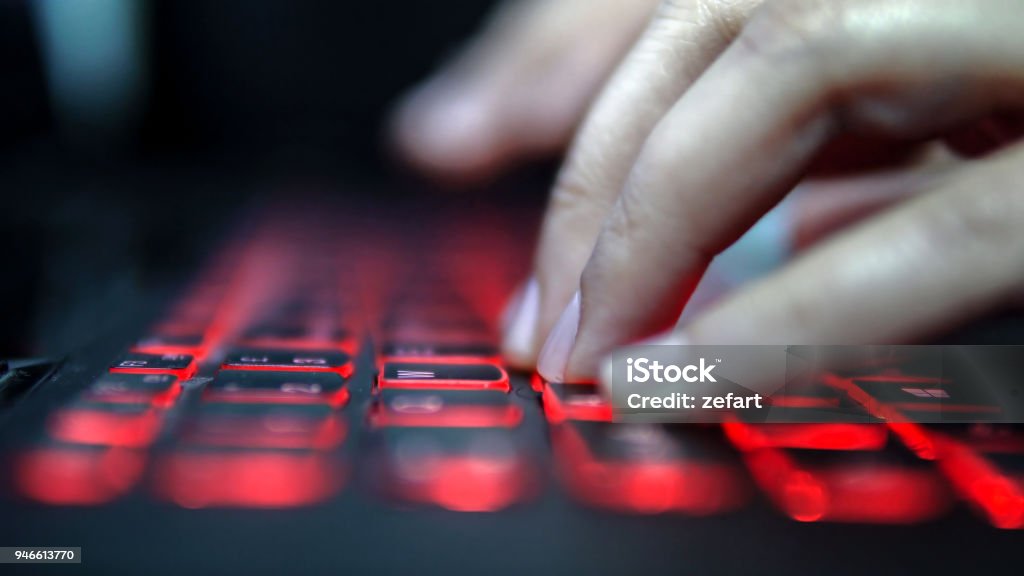 Teenage Hacker Girl Attacks Corporate Servers in Dark, Typing on Red Lit Laptop Keyboard. Room is Dark Computer Hacker Stock Photo