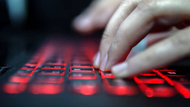 Teenage Hacker Girl Attacks Corporate Servers in Dark, Typing on Red Lit Laptop Keyboard. Room is Dark stock photo