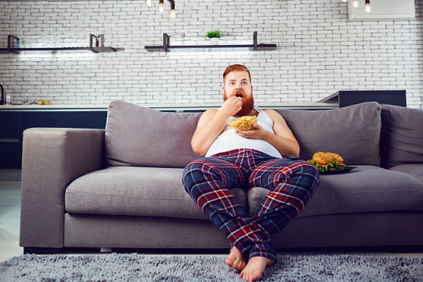 hombre divertido gruesa en pijama comiendo una hamburguesa sentado en el sofá - hombre feo fotografías e imágenes de stock