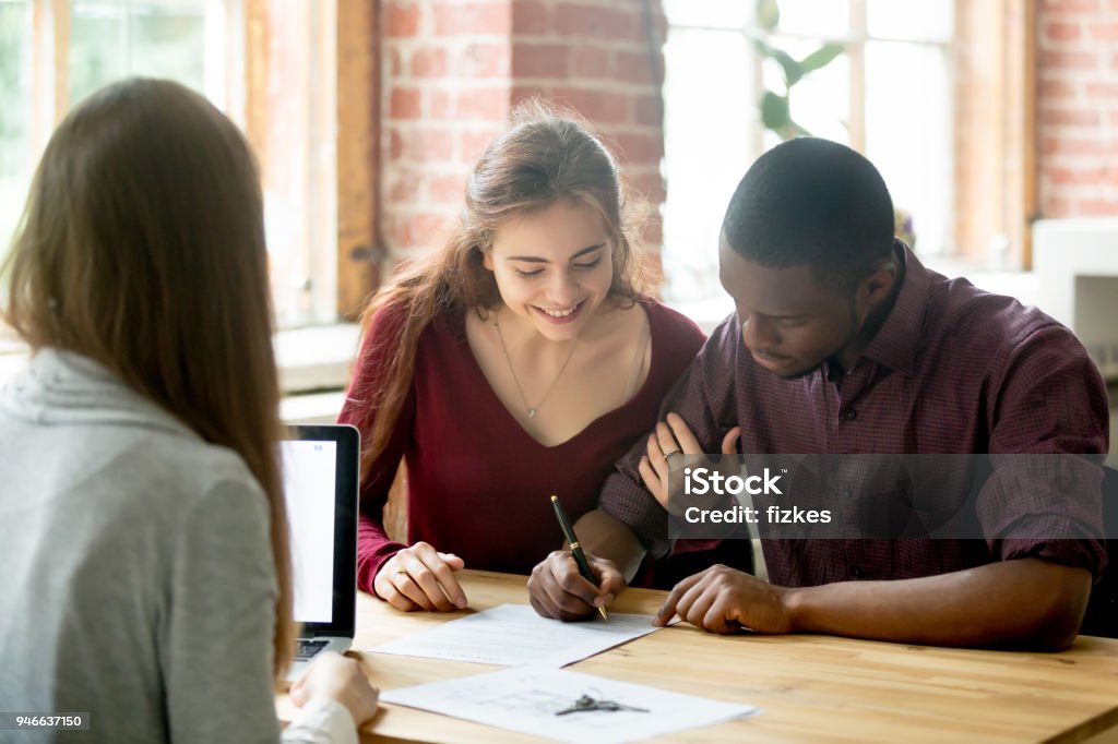 Multiracial couple signing deal from real estate agent Smiling wife watching african american husband signing purchase deal from female real estate agent. Multiracial diverse couple buying first home together, making loan, legalizing mortgage. Signing Stock Photo