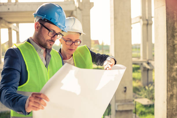 Young male and female architects or business partners looking at floor plans on a construction site Young female and male engineers or business partners at construction site, working together on building's blueprint. Architecture and teamwork concept. civil engineering stock pictures, royalty-free photos & images