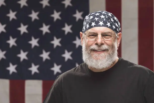 Bearded veteran standing in front of an American Flag wearing a stars and stripes skull cap.