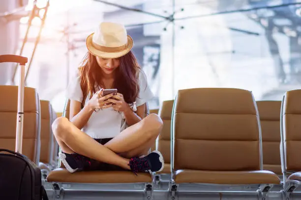 Photo of Asian woman teenager using smartphone at airport terminal sitting with luggage suitcase and backpack for travel in vacation summer relaxing waiting flight transport