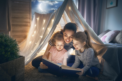 Family bedtime. Mom and children daughters are reading a book in tent. Pretty young mother and lovely girls having fun in children room.
