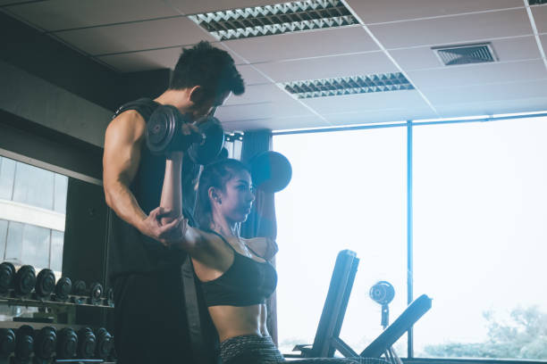 istock Personal training men help women's arms to pull up dumbbell for exercise at indoor gym. 946861708