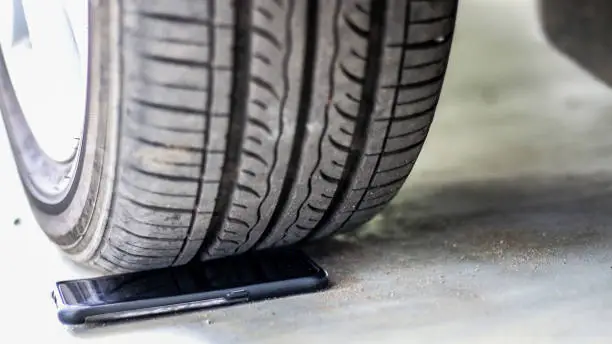 A black smart phone with a housing is on a cement floor just about to run over by a car tyre.