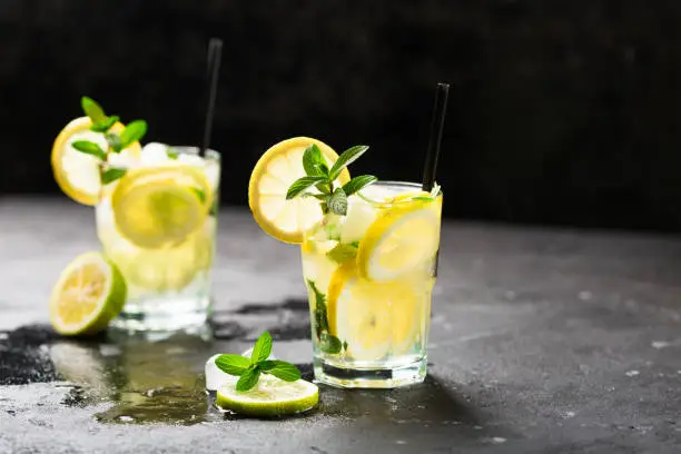 Lemon fruit lime slices caipirinha from Brazil, lemonade mint ice cubes in cold glasses on dark background, alcoholic mojito cockail ice tea green mint leaves, brown sugar spoon, copy space