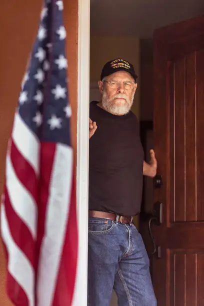 Bearded Vietnam veteran standing in his doorway with an American Flag in the foreground.