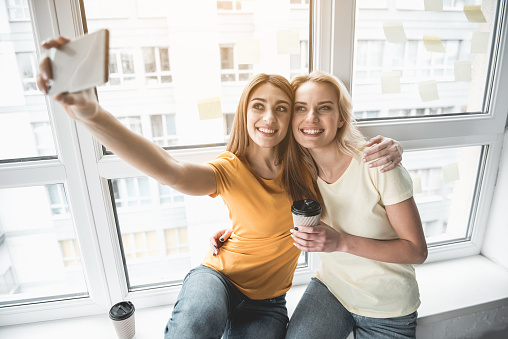 Portrait of two young women sitting with paper cups and photographing themselves. They are smiling