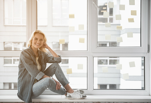Full length portrait of cheerful girl looking at camera and laughing while reposing on windowsill. Copy space in right side