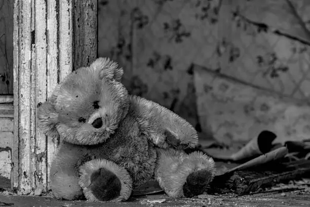 A dirty teddy bear leaning against a door frame in an old, abandoned house. Debris and assorted wallpaper peeling from the wall in the background. Black and white.