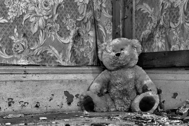 A dirty teddy bear sitting in the corner in an old, abandoned house. Debris on the floor and wallpaper peeling from the wall. Black and white.