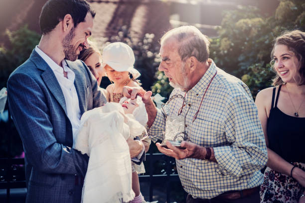 Outdoors baby baptism with family and celebrant. Baptism are not always held in church. Some families prefer to celebrate in the confort of their own backyard, with a family member as the celebrant. Father is holding the baby with the traditional robe. Godmother appearing in the picture. Horizontal mid-length outdoors shot. baptism stock pictures, royalty-free photos & images