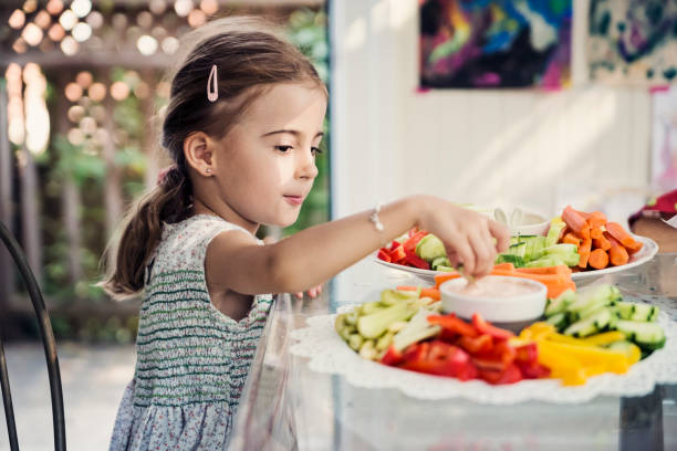 Cute little girl eating raw vegetables at a buffet. Cute adorable little girl in flowers cotton dresses eating raw vegetable at a buffet in family reunion. She is dipping some in sauce. Horizontal waist up indoors shot with copy space. dipping stock pictures, royalty-free photos & images
