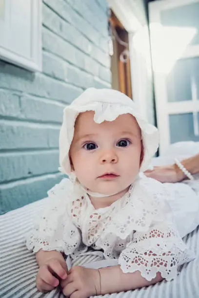 Adorable and expressive baby girl lying on her stomach on the changing table, looking at the camera. She is wearing a white lace dress and hat. Mother’s hand holding her. Vertical indoors full length shot with copy space.