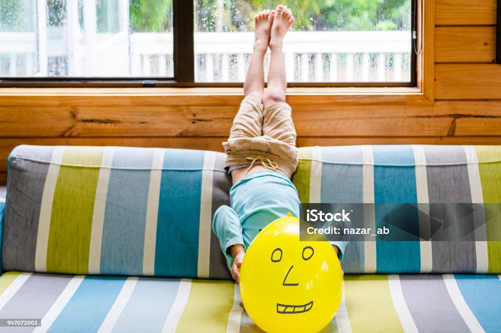 Kid lying upside down on sofa at home and covering face with balloon. Kid holding Emoji face balloons and playing at home, in Auckland, New Zealand. 4-5 Years Stock Photo
