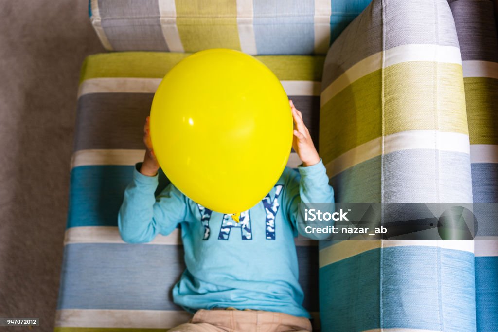 Top view of kid lying on sofa and covering his face with balloon. Kid holding Emoji face balloons and playing at home, in Auckland, New Zealand. 4-5 Years Stock Photo