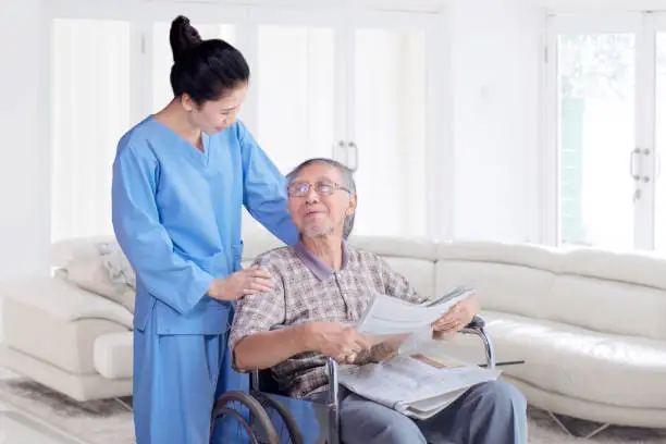 Elderly man sitting on wheelchair while talking with his nurse in the living room at home