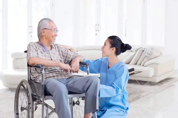Happy Asian elderly man talking and smiling with his nurse while sitting on wheelchair in the living room at home