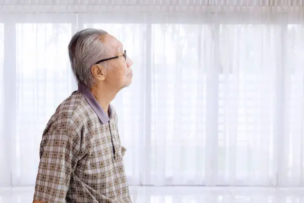 Lonely Asian elderly man wearing glasses and standing near the window while daydreaming at nursing home