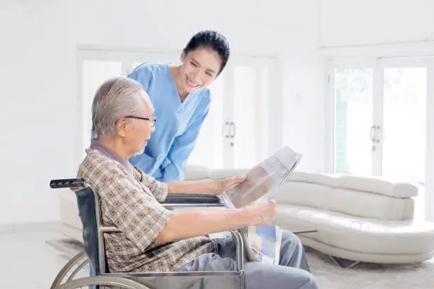 Asian female nurse talking and smiling at an old man on wheelchair in the living room at home