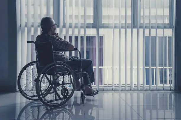 Lonely old man sitting on wheelchair while staring out of a window as he longs for his freedom and friends