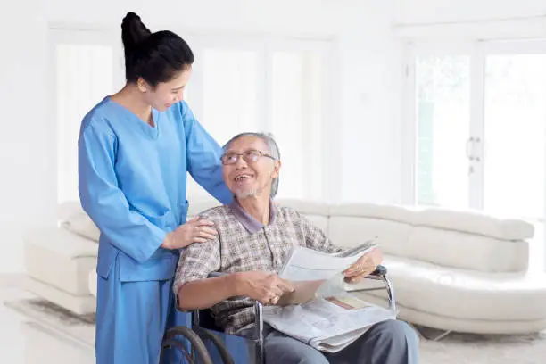 Old asian man sitting on wheelchair and smiling happy while talking with his nurse in the living room at home