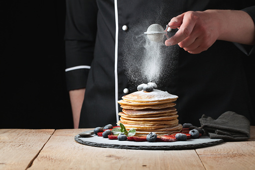 Serving pancakes with powdered sugar and berries. Chef man hand. Beautiful food still life. slightly toned image, dark black background with text area. Horizontal view.