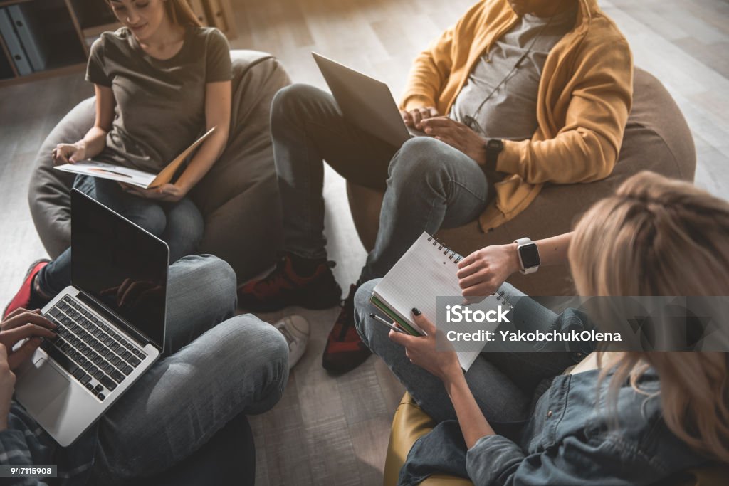 Young command working on common project Calm guys and girls taking seats on beanbags and doing their work. Men using notebooks. Woman checking time on smartwatch Flexibility Stock Photo