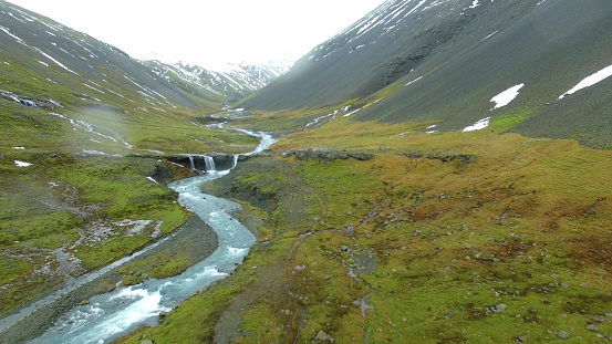 In front there is meadow surrounded by mountains. By it's left side, there is brook, and far in front there is small waterfall. Mountains are half covered by snow. Grass round brook is whole green. Iceland is perfect place for explorers and adventure people.
