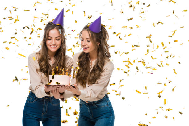 smiling twins in birthday caps looking at birthday cake under falling confetti isolated on white - twin falls imagens e fotografias de stock