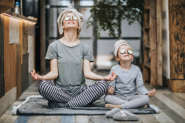 Relaxed mother and daughter exercising Yoga in the morning at home. Mother and little girl taking care of their bodies in the morning while doing Yoga meditation exercises at home. Their hair is wrapped in towels while cucumber is on her eyes. cross legged photos stock pictures, royalty-free photos & images