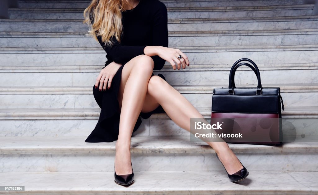 Fashion model sitting in black dress with big bag high heel shoes Fashion model in black dress with big leather red handbag in high heel shoes sitting on the white stair Women Stock Photo