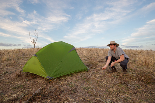A series of photos showing a man in his 30's setting up the fly on his tent, followed by pegging the corners and guide ropes. The scene is shot in an open field with sweeping views and stunning afternoon lighting.