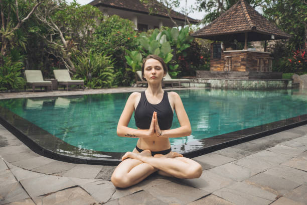 Woman sitting in lotus pose  near the swimming pool on villa Young Caucasian woman sitting in lotus pose  near the swimming pool on villa women exercising swimming pool young women stock pictures, royalty-free photos & images