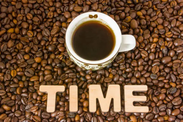 Visualization of the concept or action Time Coffee. Word time, which is lined with large, 3D letters, lies in scattered roasted beans coffee near small mug, which is brewed flavored drink