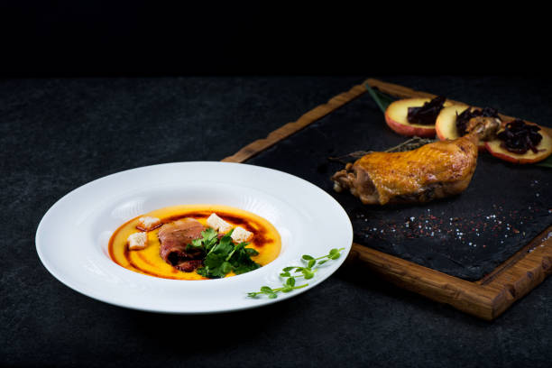 An appetizing pumpkin soup is served in a white plate in a restaurant. Duck confit with baked apple