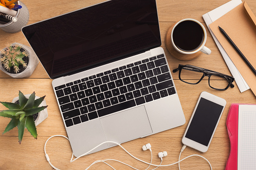 Male workplace with laptop and smartphone with blank screen for advertisement, coffee cup and glasses on wooden brown background, top view, copy space.