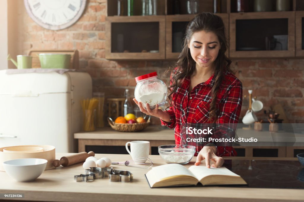 Happy young woman baking in loft kitchen Happy young woman baking pie in loft kitchen at home, using book with recipe. Mockup, copy space Cooking Stock Photo