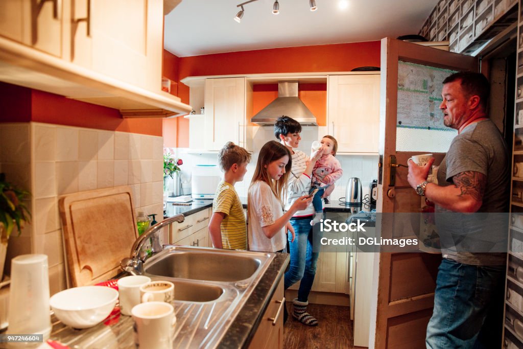 Family Life in the Kitchen A whole family are busy in the kitchen of their home. The mother is feeding the baby while the father does the dishes. The older girl is using a smartphone and her brother is watching the baby get fed. Crowded Stock Photo