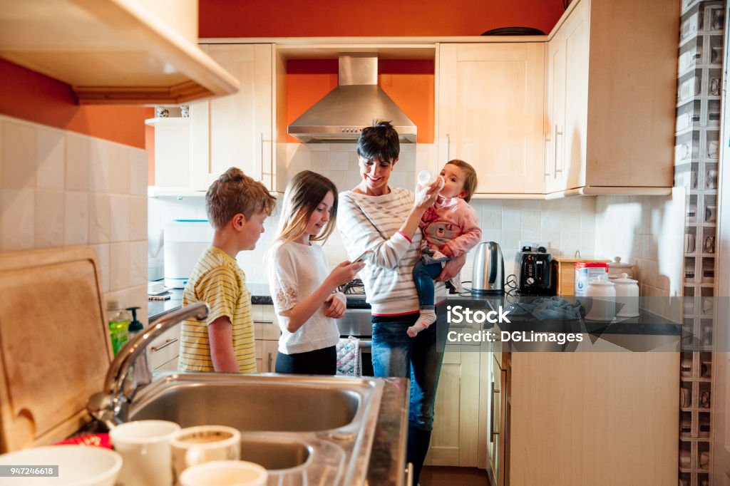 Family Socialising in the Kitchen Multitasking mum is tending to all of her children at once in the kitchen of their home. She is feeding the baby a bottle while talking to her eldest children and looking at their smart phone. Multi-Tasking Stock Photo