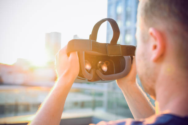 istock VR Goggles. Man ready to wear virtual reality goggles. The vr headset design is generic and no logos. Backward light, selective focus, close up. Sunset city landscape. 947252490