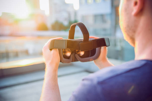 istock VR Goggles. Man ready to wear virtual reality goggles. The vr headset design is generic and no logos. Backward light, selective focus, close up. Sunset city landscape. 947252502