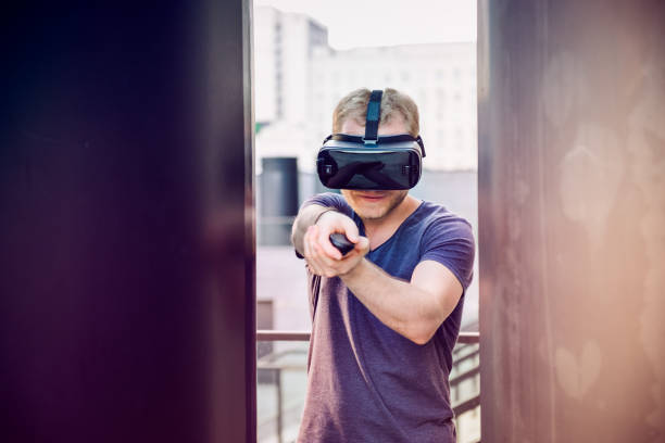 istock Young man playing shooting games in virtual reality headset on the urban building background outdoors. Technology, innovation, cyberspace and gaming. Selective focus. 947252516