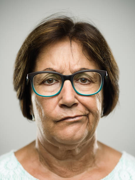 Real displeased senior woman portrait Close up portrait of hispanic mature woman with bored expression against white background. Vertical shot of real senior woman in studio. Short brown hair and modern glasses. Photography from a DSLR camera. Sharp focus on eyes. grimacing photos stock pictures, royalty-free photos & images