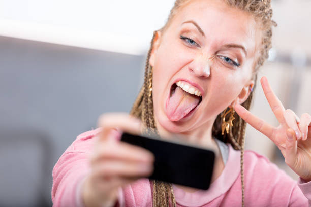 Young woman pulling a goofy face for a selfie Young woman pulling a goofy face for a selfie sticking out her tongue and making a V-sign gesture as she poses for her mobile phone camera derogatory stock pictures, royalty-free photos & images