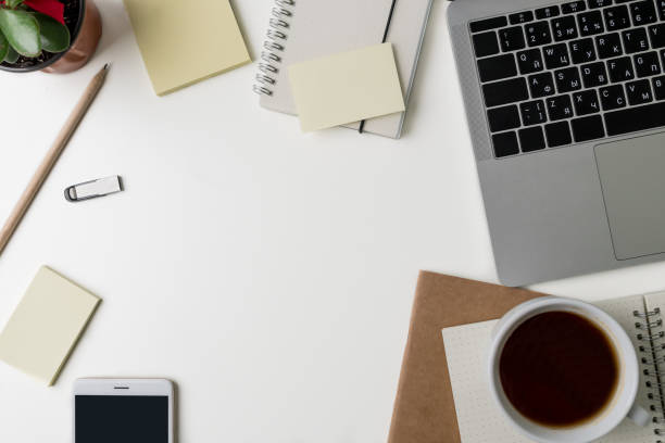 istock Top view of office workplace. White desk with copy space. Flat lay view on table with laptop, glasses, pen, notebook, green flower, flash memory card, coffee and stickers. Creative designer concept. 947279214