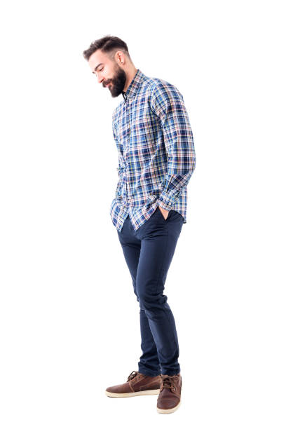 Shy handsome bearded young man in plaid shirt with hands in pockets smiling and looking down Shy handsome bearded young man in plaid shirt with hands in pockets smiling and looking down. Full body isolated on white background. hands in pockets stock pictures, royalty-free photos & images