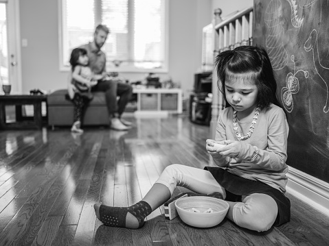 Young mixed race family with two daughters. Having a play time together. Interior of living room, day time. Daughter of 4 -6 years old is drawing pictures with chalk on black board in the living room. Father with other daughter playing in the background.